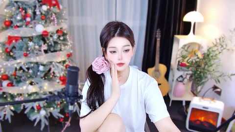 Media: A video of a young Asian woman with fair skin and long dark hair, wearing a white T-shirt, seated in front of a decorated Christmas tree and a lit fireplace in a cozy room.