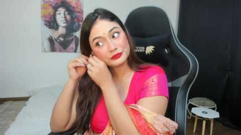 Media: Video of a young South Asian woman with long dark hair, wearing a bright pink blouse, adjusting her hair in a black gaming chair; colorful wall art and drum set in the background.