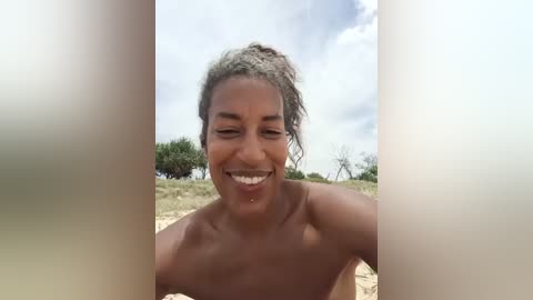 Media: Video of a joyful, topless Black woman with curly hair, smiling widely outdoors with a clear sky background.