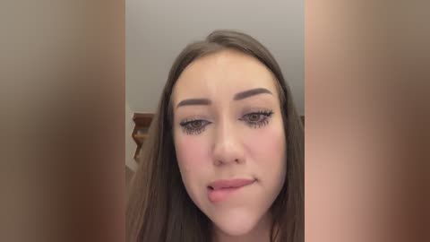 Media: A video of a young Caucasian woman with long brown hair, fair skin, and makeup featuring bold eyeliner and mascara, sticking out her tongue playfully. Background is a blurred indoor setting with a wooden staircase.