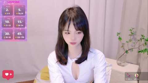 Media: Video of a young East Asian woman with shoulder-length dark hair, wearing a white blouse with a keyhole neckline, sitting on a bed. Background features a minimalist room with a vase of green branches and a pink digital display showing her stats.