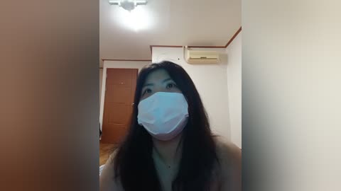 Media: Video of an Asian woman with long black hair, wearing a blue face mask, standing in a small, dimly-lit room with beige walls, a wooden door, and a ceiling light.