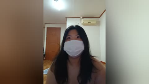 Media: Video of an Asian woman with long black hair, wearing a white surgical mask and light-colored top, standing in a simple, sparsely furnished room with wooden flooring, white walls, and a wooden door.