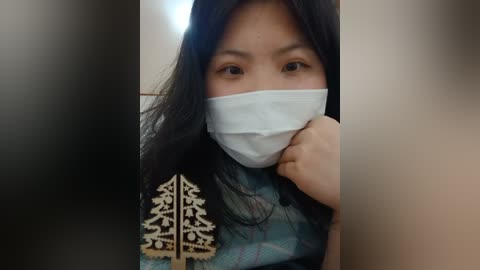 Media: Video of a young Asian woman with long black hair, wearing a white surgical mask, a blue shirt with a golden tree pattern, and a hand near her face, partially obscured by a blurred background.