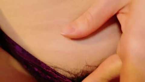 Media: Video of a close-up of a person's hand gently pressing against their own lower abdomen, revealing a patch of pubic hair. The skin is light, and the background shows a hint of purple lace underwear.