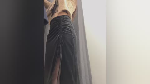 A video of a woman with light skin, wearing a beige cropped sweater and a high-waisted black skirt with a slit, standing in a dimly lit room with gray walls and a white door.