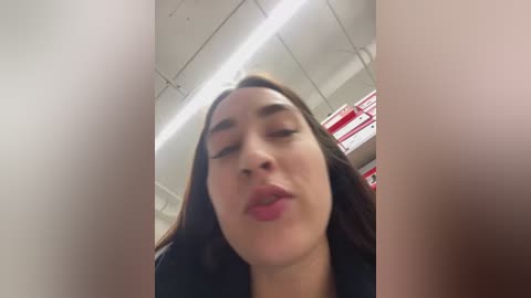 Video of a young woman with light skin, long dark hair, and closed eyes, puckering her lips, taken from a low angle. The background shows a fluorescent-lit ceiling and a red and white striped object, possibly a sign or banner.