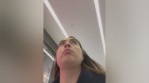 Video of a woman with long brown hair, fair skin, and wearing a black jacket, gazing upwards in an indoor setting with white ceiling and fluorescent lights.