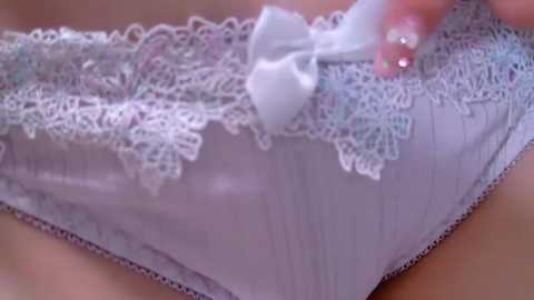 Media: Video of a woman's waist, showing delicate white lace panties with intricate floral patterns and a small white bow. Her hand with pink nails is adjusting the panties.