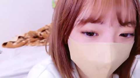 Media: A close-up video of a young woman with light skin and shoulder-length, straight, reddish-brown hair. She wears a beige surgical mask, and her large, dark eyes gaze forward. The background shows a blurred, light-colored bed with a brown blanket.