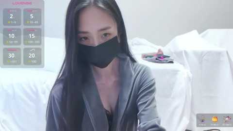 Media: Video of an East Asian woman with long black hair, wearing a black face mask, gray robe, and revealing a black bra underneath. Background shows a bed and a digital health monitor.