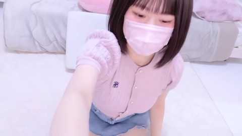 Media: Video of a young Asian woman with a pale complexion and straight black hair, wearing a light pink mask, short-sleeve shirt, and denim shorts, sitting on a white floor.