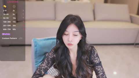 Media: A video of an East Asian woman with long, wavy black hair, wearing a black lace dress, seated in a modern living room with beige sofa and blue cushions.