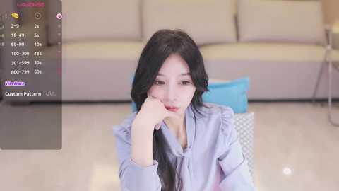 Media: Video of an East Asian woman with long black hair, wearing a light blue blouse, sitting on a white chair in a modern, minimalist living room.