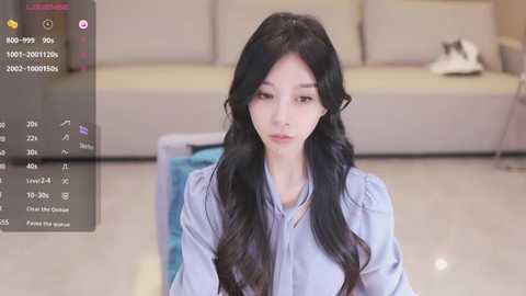 Media: Video of a young Asian woman with long black hair, wearing a light blue blouse, sitting on a light-colored sofa in a modern living room with a beige background.