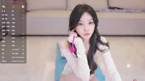 Media: Video of a young East Asian woman with long black hair, wearing a white lace sweater, sitting on a blue mat, talking on a pink phone. Background shows a beige sofa and a blurred purple object.