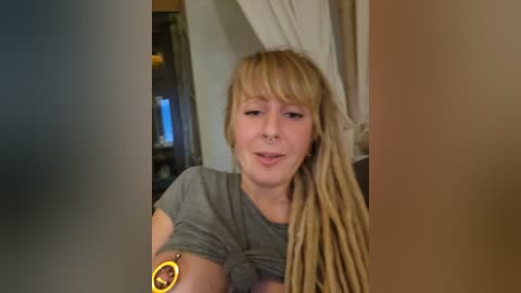 Media: A video of a young woman with long, blonde dreadlocks, wearing a gray t-shirt and a nose ring, smiling, indoors, with a blurred background.