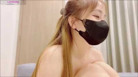 Media: Video of a light-skinned Asian woman with long, straight brown hair, wearing a black face mask, topless, looking to the right in a neutral indoor setting.