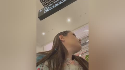 Media: Video of a young woman with long, straight brown hair, wearing a floral-patterned top, standing in a brightly lit, modern indoor setting with a ceiling-mounted sign.