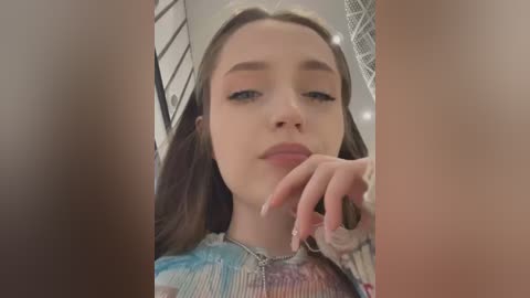 Media: Video of a young woman with fair skin, brown hair, and light makeup, wearing a colorful tie-dye top. She gazes directly at the camera, fingers lightly touching her lips.