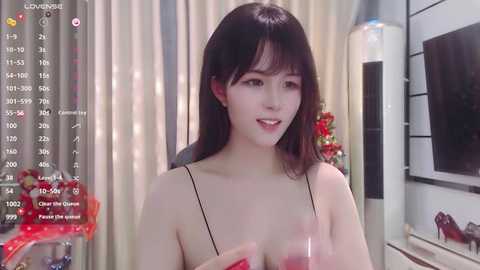 Media: Video of an East Asian woman with straight black hair and fair skin, wearing a black spaghetti strap top, holding a glass of red wine, indoors with a festive Christmas tree in the background.