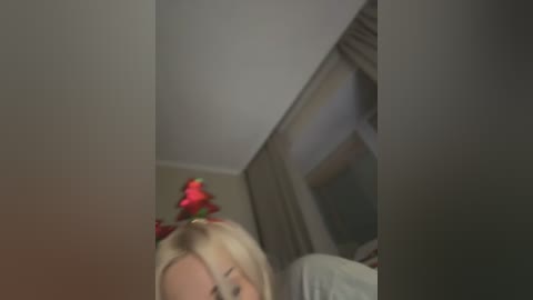 Media: A blurred video shows a blonde woman in a red ribbon headband, partially visible. Background features a window with closed beige curtains and a plain wall.