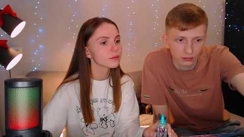 Media: Video of two young people, a girl with long brown hair and a boy with short blonde hair, watching TV, both wearing casual clothes. Background features a festive, twinkling light display and a colorful Christmas tree.