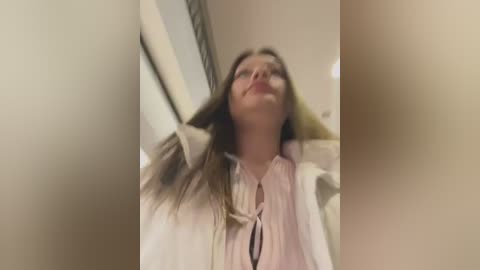 Media: Video of a young Caucasian woman with long brown hair, wearing a white blouse and pink vest, leaning against a doorframe in a dimly lit room.