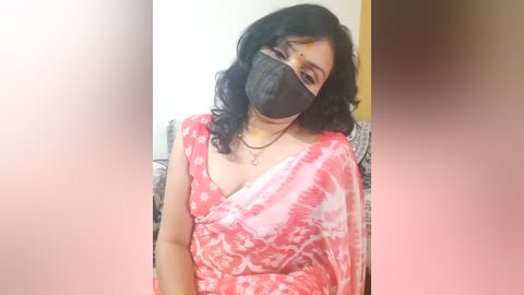 Media: A video of a woman with dark curly hair, wearing a red saree with white floral patterns, a black face mask, and a necklace. The background is blurred, showing a beige wall and a patterned cushion.