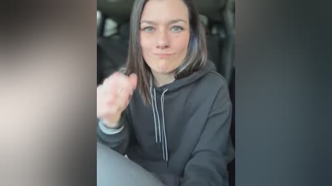 A video of a young woman with straight brown hair, wearing a black hoodie, smiling and making a thumbs-up gesture while seated in a car.