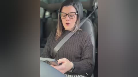 Media: Video of a woman in glasses, wearing a knit sweater, seated in a car, with a seatbelt across her chest, holding a tablet, looking at it with an expression of surprise or concern.