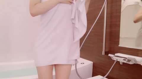 Media: Video of a person in a white towel, standing in a modern bathroom with wooden paneling, white walls, and a bathtub, holding a phone.