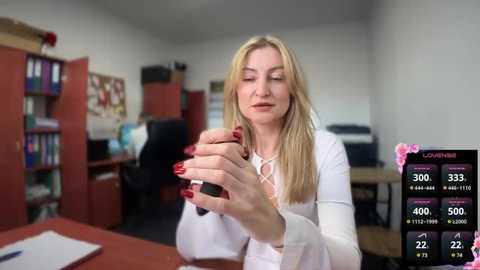 Media: Video of a blonde woman in a white top, sitting at a desk in a cluttered office, holding a black pen, with a live poker game screen in the background.