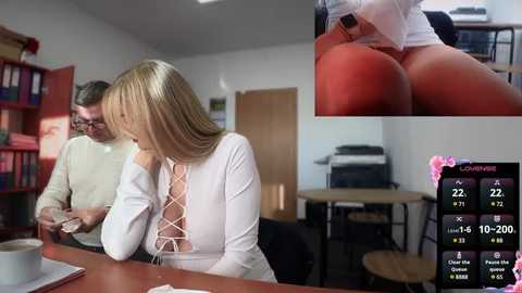 Media: A video shows a blonde woman in a white blouse, sitting at a desk with a man beside her. An inset image highlights her bare buttocks. The room is a modern office with bookshelves and a calendar displaying dates.