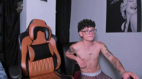 Media: Video of a shirtless young man with curly hair, wearing glasses, tattoos, and plaid pajama pants, sitting in a tan and black gaming chair, against a gray wall with Marilyn Monroe poster.