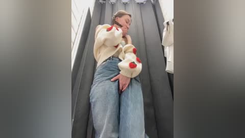 Media: Video of a woman wearing a cream sweater with red plush bear ears and blue jeans, sitting on a moving escalator, hands on thighs, in a shopping mall.