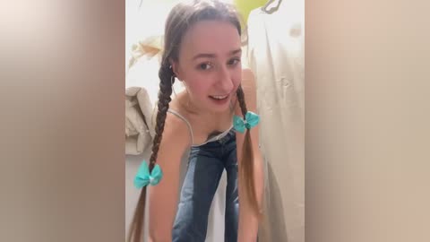 Media: Video of a young, smiling Caucasian woman with long brown hair in twin braids, wearing a white tank top and blue jeans, standing in a small, cluttered room with beige walls and clothes hanging.