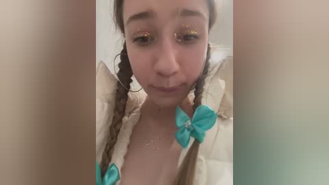 Media: Video of a young woman with light skin, braided hair, wearing a cream-colored jacket with turquoise bow accents and gold glitter eyeshadow, looking down.