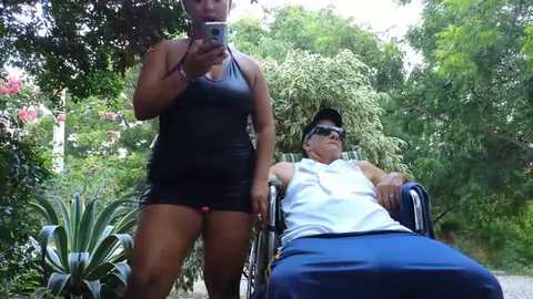 Media: Video of an African American woman pushing a white man in a wheelchair along a garden path. She wears a black sleeveless top and shorts, he in a white tank top and blue shorts. Lush greenery surrounds them.