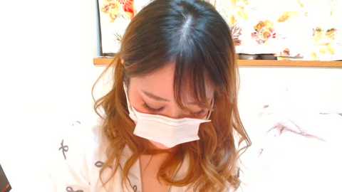 Media: Video of an Asian woman with long, wavy brown hair, wearing a white surgical mask, seated indoors with a floral-patterned wall in the background.