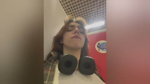 Media: A video of a young person with shoulder-length brown hair, wearing headphones, standing in a tiled bathroom with a red wall.