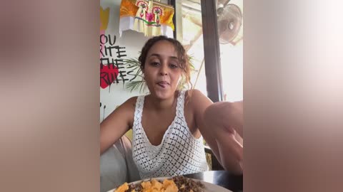 Media: Video of a young Latina woman with medium skin tone and brown hair in a bun, wearing a white crochet top, eating pizza at a sunny outdoor caf\u00e9.