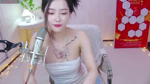 Media: Video of a fair-skinned Asian woman with dark hair, wearing a white strapless dress and jewelry, singing into a microphone in a cozy room with a plaid blanket, a plant, and a red poster.