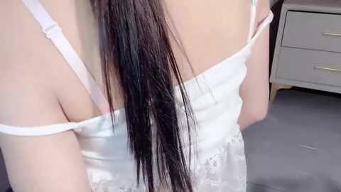 Media: Video of a woman's bare back with long, straight, dark brown hair in a ponytail, wearing a white, off-shoulder lace dress. Background features a white dresser and grey carpet.