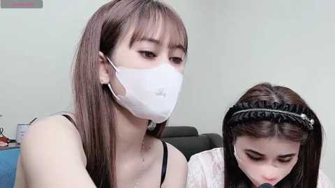 Media: Video of two East Asian women, one wearing a white face mask and black tank top, the other in a white headband, both with long hair, sitting closely in a modern, minimalistic room.