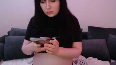 Media: Video of a young woman with pale skin and long black hair, wearing a black t-shirt, holding a smartphone, sitting on a dark gray couch in a dimly lit room.