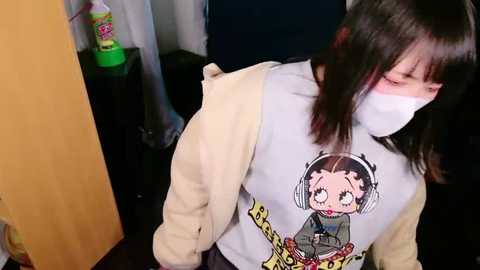 Media: Video of an East Asian woman with shoulder-length black hair, wearing a white face mask, light sweater, and a graphic tee featuring a cartoon girl with headphones, standing in a dimly lit room with a green plastic toy and a wooden door.