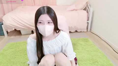 Media: A video of an Asian woman with long black hair and light skin wearing a white off-shoulder blouse, white mask, and red plaid skirt, sitting on a green rug in a pink bedroom with a bed.