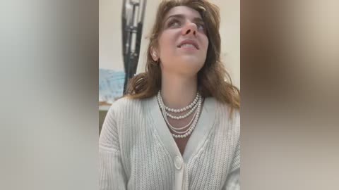 Media: Video of a fair-skinned woman with wavy brown hair, wearing a light gray cardigan with multiple pearl necklaces, looking upward. Background features a blurred, neutral-toned room with a decorative lamp.