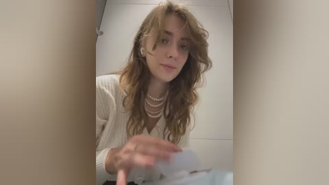 Media: Video of a young Caucasian woman with long, wavy blonde hair, wearing a white cardigan and pearls, sitting in a bathroom, holding a magazine.